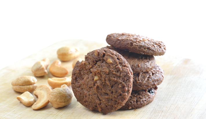 fotografia em tons de branco, marrom e bege de uma bancada bege vista de frente, ao centro biscoitos de chocolate e ao lado castanhas de caju