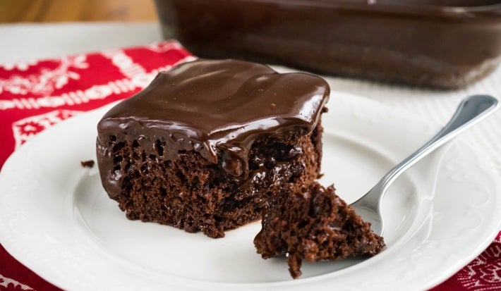 Fotografia de uma fatia de bolo de chocolate em um prato branco, com ganache brilhante e cremosa. Um garfo sugere que a fatia está pronta para ser saboreada.