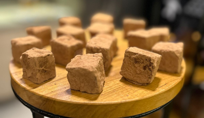 Fotografia em tons de marrom com uma bancada de madeira ao centro. Em cima da bancada existem vários quadrados de chocolate cobertos com chocolate em pó