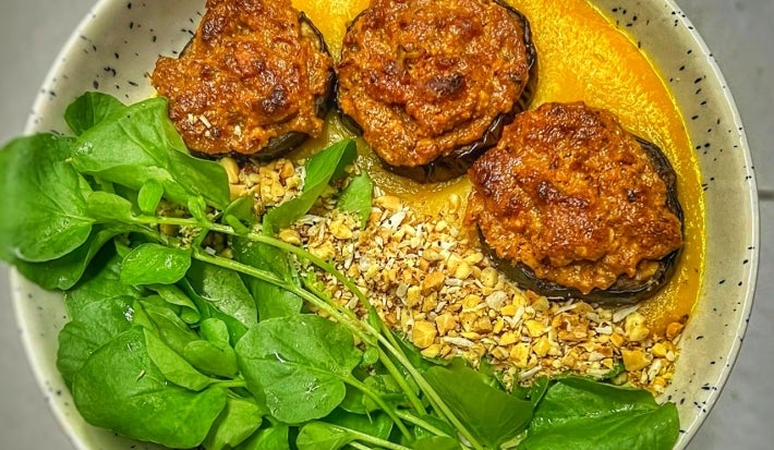 Fotografia vista de cima em tons claros de um prato claro de vidro com berinjela, queijo, molho de tomate, agrião e farofa.