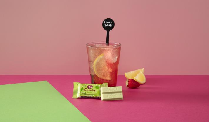 Foto da receita de Soda Italiana Pink Lemonade. Observa-se um fundo rosa e verde com um copo alto ao centro, servido cheio de gelo com uma rodela de limão siciliano dentro. À frente, um KIT KAT Limão.