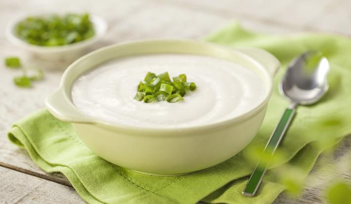 Fotografia em tons de verde em uma bancada de madeira com um guardanapo de pano verde ao centro com uma tigelinha branca e o creme de couve-flor dentro enfeitado com cebolinhas verdes. Ao lado, uma colher e ao fundo um potinho com cebolinhas verdes.