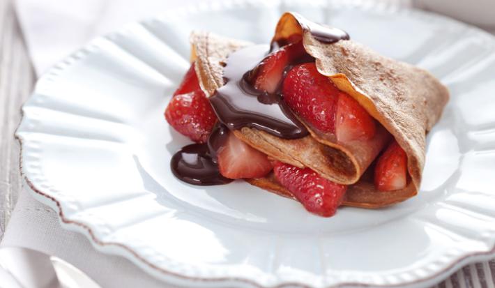 fotografia em tons de branco, marrom e vermelho de uma bancada cinza vista de frente, contém um prato branco redondo com um Crepe de Chocolate com Morangos e ao lado uma colher para servir.