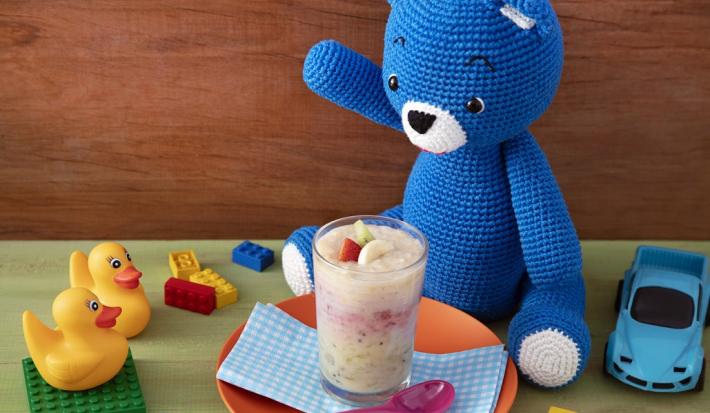 Fotografia em tons de branco em uma bancada de madeira de cor verde. Ao centro, um prato laranja contendo um pano quadriculado azul em cima com o smoothie. Ao lado, está o ursinho azul e ao redor, há carrinhos, e brinquedos espalhados.