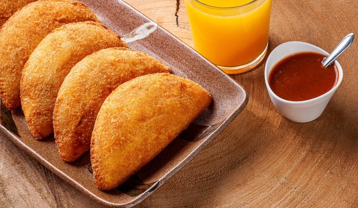 Fotografia de cinco risoles de presunto e queijo sobrepostos em um recipiente retangular marrom escuro sobre uma mesa de madeira. Ao lado dos salgados tem suco de laranja no copo de vidro e um molho vermelho em um recipiente branco pequeno.