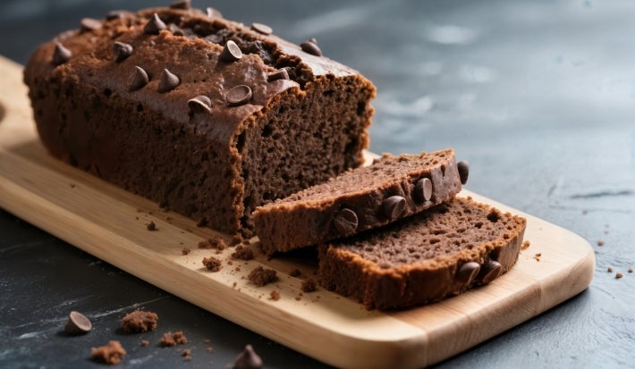 Bolo de chocolate recém-assado em uma tábua clara, coberto com gotas de chocolate. Duas fatias cortadas mostram a textura úmida. A apresentação casual é realçada por migalhas e um fundo escuro, destacando o bolo apetitoso.