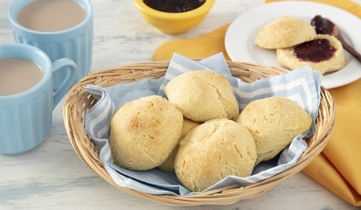 Foto da receita de Pão de Leite em Pó. Observa-se uma cestinha com um guardanapo azul e branco com 5 pãezinhos dentro. Ao lado esquerdo, duas canecas com café com leite. Do direito, um pratinho com um pão cortado ao meio recheado de geleia.