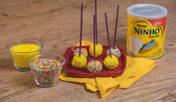 Fotografia em tons de amarelo e vermelho em uma bancada de madeira clara, um pano amarelo e um prato vermelho com alguns pirulitos de ninho com granulado colorido em cima dele. Ao lado, potinhos com os granulados coloridos e a lata de Ninho em Pó.