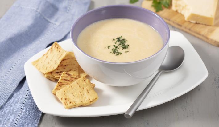 Fotografia em tons de cinza, branco e azul de uma bancada cinza com um prato branco retangular, sobre ele um recipiente redondo branco com o creme de queijo e torradinhas. Ao lado um paninho azul. Ao fundo uma tábua com um pedaço de queijo e salsinha.