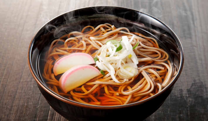 Fotografia mostra um macarrão japonês sendo segurado por hashi, ao fundo um bowl escuro com macarrão dentro.