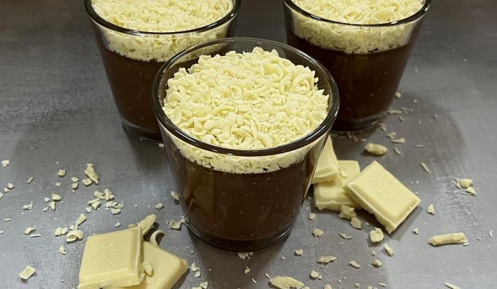Foto da receita de trufa cremosa servida em três porções em copos de vidro baixos sobre uma mesa acinzentada com pedaços e raspas de chocolate branco rodeando os copos como decoração