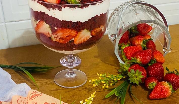 Foto em tons de vermelho da receita de red velvet na taça servida em uma graned porção em uma taça de vidro alta em cima de uma mesa de madeira com um pote de vidro com morangos ao lado
