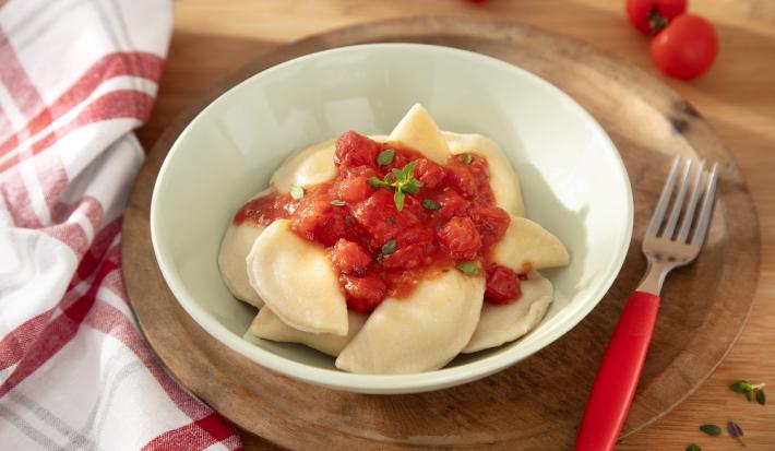 Foto da receita de Pierogi (massa polonesa). Observa-se um bowl verde claro com a massa dentro, servida de molho de tomate, sobre uma tábua de madeira escura com um garfo ao lado direito.