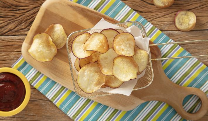Imagem vista de cima de chips de batata-doce sobre uma tábua de madeira com um potinho amarelo com ketchup ao lado.