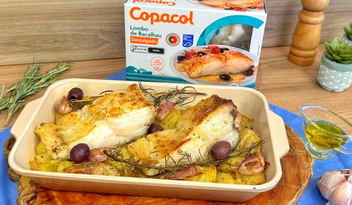 Fotografia em tons de azul e castanho com uma travessa ao centro. Dentro da travessa existem dois lombos de bacalhau assados juntamente com batatas de tamanho pequeno. Ao fundo existe uma embalagem de bacalhau congelado da marca Copacol.
