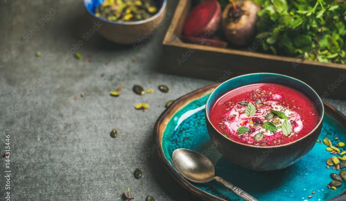 Fotografia de um recipiente azul com uma sopa de beterraba com folhas de sálvia, sementes e um creme branco por cima. Embaixo da sopa, um prato azul de tom mais claro, com uma colher de sopa ao lado. Ao fundo, beterrabas e sementes sobre uma mesa cinza.