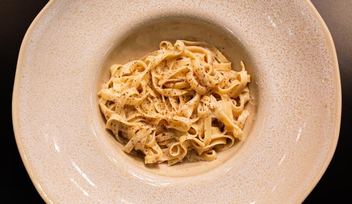 Fotografia em tons de cinza e preto com um prato cinza ao centro. Em cima do prato existe uma receita de cacio e pepe feita com macarrão tagliatelle e finalizado com pimenta preta moída