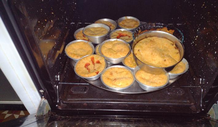 Fotografia de várias forminhas de alumínio com tortinhas de atum dentro de uma forma maior e redonda. As forminhas estão dentro do forno.