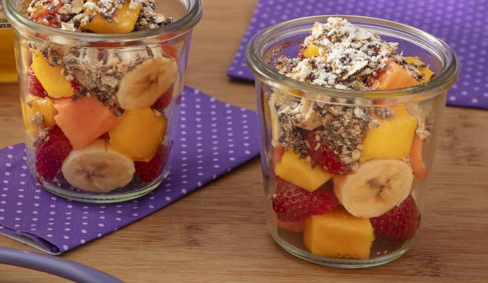 Fotografia de dois copos de vidro com frutas e granola sobre um paninho roxos com bolinhas brancas, sobre uma mesa de madeira.