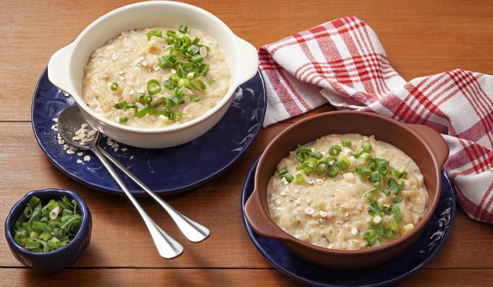 fotografia em tons de azul e branco de uma bancada marrom vista de cima, contém dois pratos redondos azuis e ambos com um recipiente redondo com alças, um cor branco e o outro marrom e ambos contém sopa cremosa com cebolinhas por cima.
