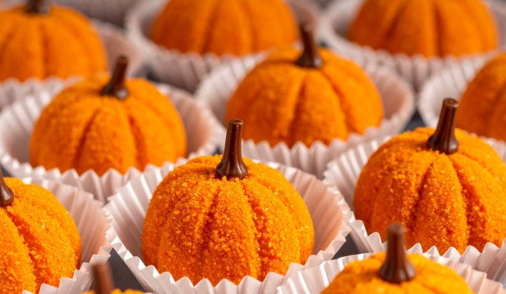 Fotografia em tons de laranja com vários brigadeiros em formato de abóbora ao centro. Os brigadeiros são feitos com abóbora cozida, passados no açúcar e finalizados com um cravo, para imitar a forma de uma abóbora.