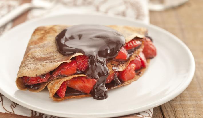 Fotografia em tons de marrom em uma bancada de madeira com um pano branco com listras marrom, um prato branco raso com o crepe recheado com morangos e chocolate.