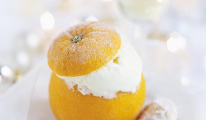 Fotografia em tons de branco com uma laranja inteira recheada de creme apoiada sobre um prato, em cima de uma mesa branca. Ao fundo, uma taça com champanhe.