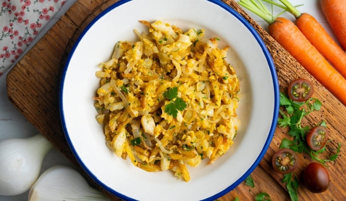 Em cima de uma mesa, prato redondo com porção de bacalhau desfiado com cebola, decorado com folha de salsa.