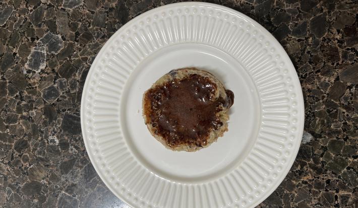 Fotografia vista de cima de um prato raso de vidro branco com um bolinho de micro-ondas com uma calda de chocolate feita de whey. O prato está sobre uma bancada escura.