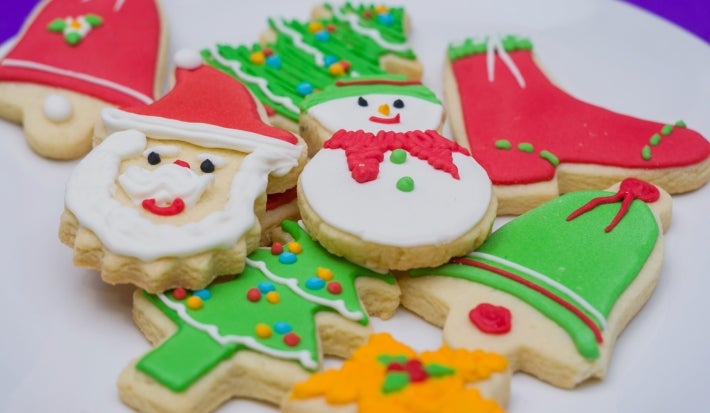 Em cima de uma mesa, prato branco com biscoitos de papai Noel, árvore de Natal e botas, decorados com glacê colorido e confeitos em forma de bolinha.