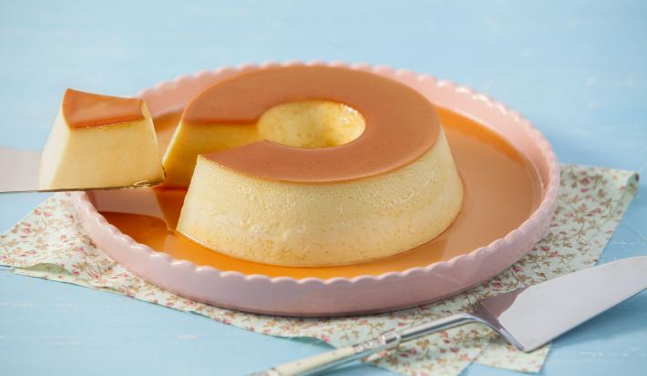 Imagem de um prato rosa claro com pudim de leite condensado com calda de caramelo e uma fatia sobre uma espátula.