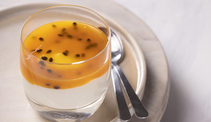 Fotografia de um creme delicado à base de maracujá e creme de leite, e por cima, uma calda de maracujá dentro de um pote de vidro pequeno. Ao lado tem duas colheres de sobremesa que estão apoiadas em um prato raso de vidro bege.