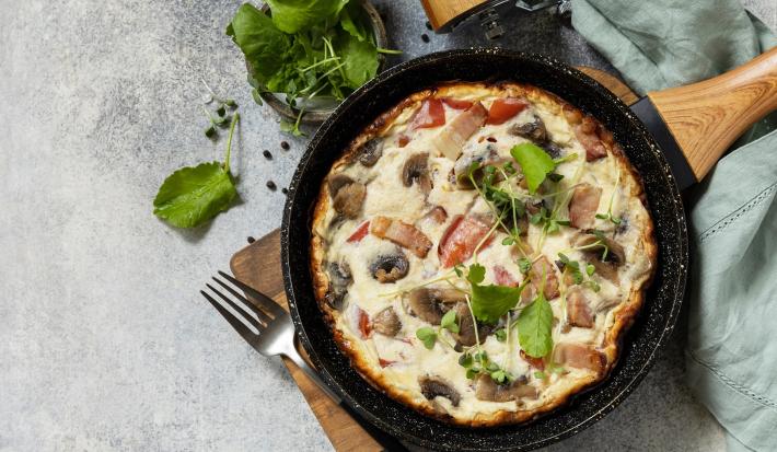 Fotografia em tons de azul claro e branco om uma frigideira ao centro. Dentro da frigideira existe um omelete feito com carne, cogumelos e algumas folhas