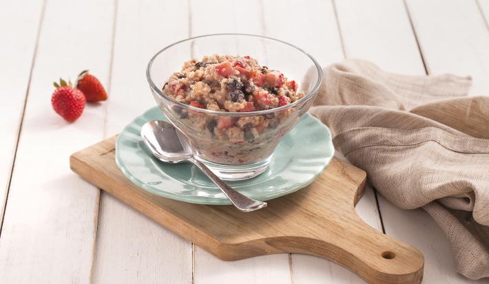 Fotografia em tons de azul em uma bancada de madeira clara, uma tábua de madeira, um paninho bege, um prato azul redondo pequeno, uma taça de vidro com o doce de quinoa com frutas secas, morango e iogurte dentro dele.