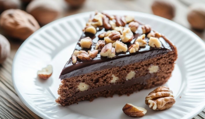 Fatia de bolo de chocolate com recheio cremoso e pedaços de nozes na massa. Cobertura lisa de ganache e finalização com nozes picadas por cima, servida em um prato branco. Um clássico com toque crocante.