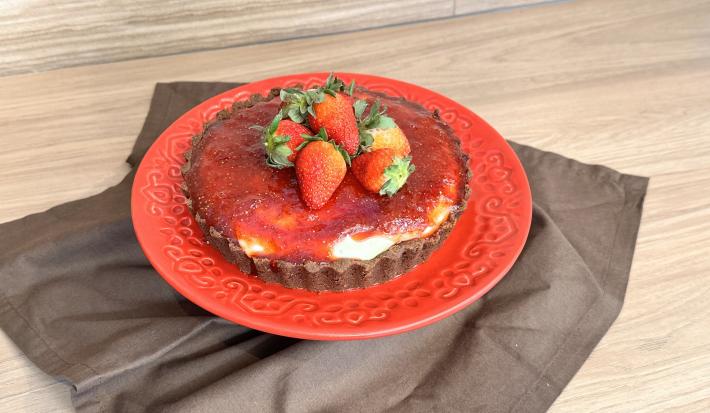 Fotografia em tons de vermelho com um prato vermelho ao centro. Em cima do prato existe um cheesecake de Galak com queijo gorgonzola, cobertura de morango e morangos inteiros