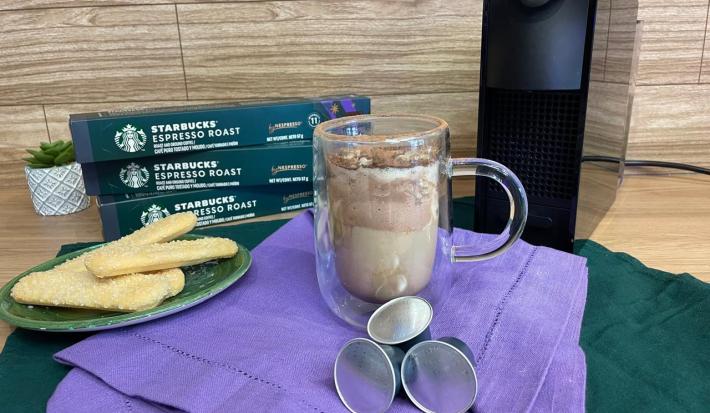 Fotografia em tons de verde e roxo em uma bancada de madeira, um paninho verde e roxo ao centro com uma xícara de vidro e o café tiramisu dentro. Ao fundo, uma máquina nespresso.