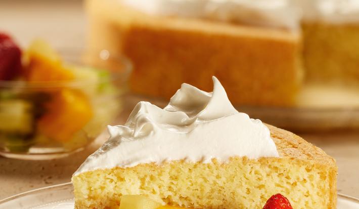 Fotografia em tons de vermelho em uma bancada com uma toalha branca com detalhes em flores vermelhas e folhas verdes. Ao centro, um prato de vidro com o pastel de tres leches com frutas.