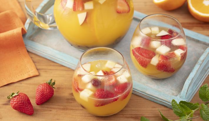 Imagem de dois copos com ponche com pedaços de frutas. Ao fundo uma jarra da bebida e ao lado morangos e folhas de hortelã.