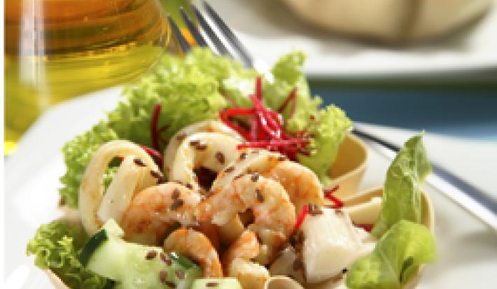 Fotografia em tons de verde em uma mesa com um vidro de azeite, um prato branco raso, um recipiente branco em formato de concha com a salada de alface, beterraba ralada, cenoura ralada e camarões dentro dele.