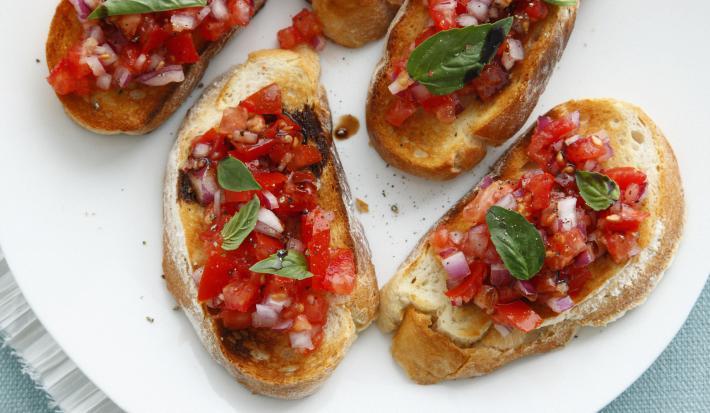 Fotografia vista de cima de cinco fatias de pão italiano torrado com uma bruschetta por cima. Os aperitivos estão sobre um prato de vidro branco, que está apoiado em um pano de mesa de cor azul.