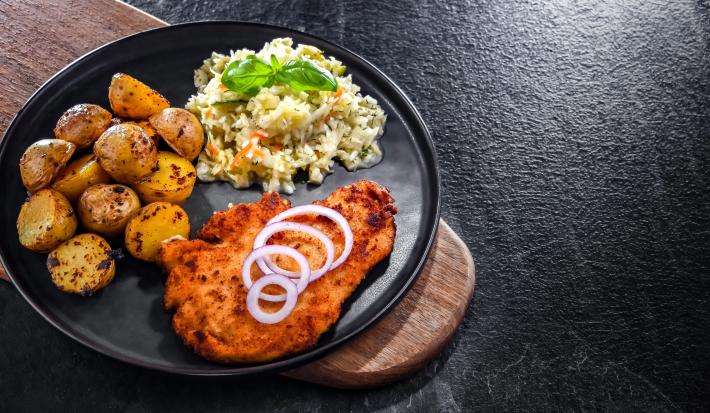 Fotografia em tons de preto com um prato preto ao centro. Em cima do prato existe uma receita de frango à milanesa acompanhada de repolho.
