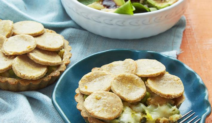 Fotografia em tons de azul em uma mesa de madeira laranja com um pano azul e um prato redondo fundo branco com salada e um prato fundo azul com a torta de massa de grão-de-bico com vegetais