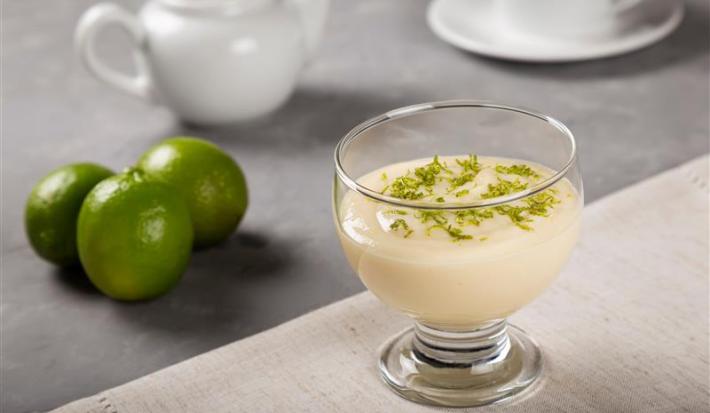 Fotografia em tons de branco com uma taça de sobremesa de vidro ao centro dentro da taça existe uma receita de mousse de limão coberta com raspas de limão.