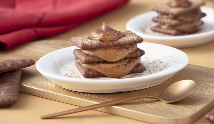 Foto de uma mesa marrom clara. Bem ao centro está uma tábua de madeira marrom clara com um prato branco contendo a receita de Tiramisù de ChocoBiscuits polvilhado com chocolate em pó. Há uma colher dourada, biscoitos espalhados e um guardanapo de pano.