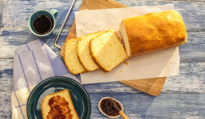 Fotografia em tons de azul e branco de uma bancada azul com uma tábua, sobre ela um paninho branco com o pão. Ao lado um paninho branco e azul, sobre ele um prato redondo azul com uma fatia de pão e geleia e uma xícara de café.
