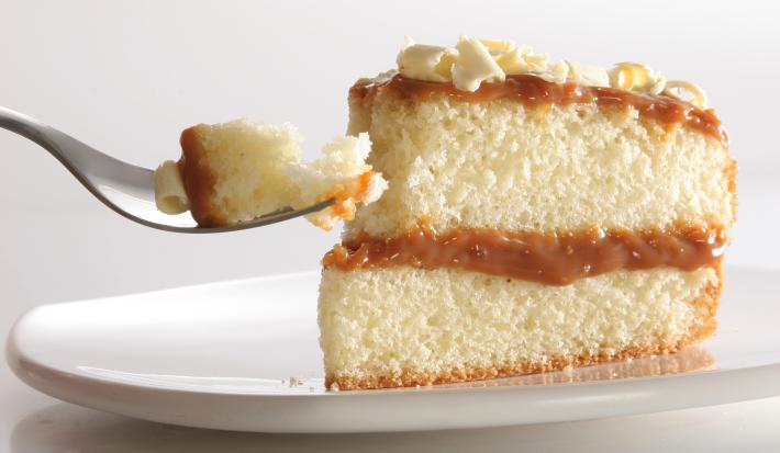Fotografia de uma fatia de bolo bem-casado com doce de leite sobre um prato raso branco apoiado em uma mesa branca.