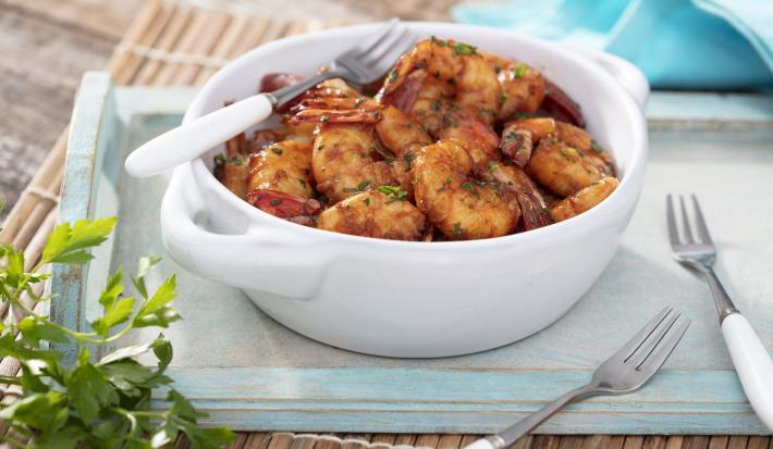 Fotografia em tons de azul em uma bancada de madeira clara, uma bandeja de madeira azul, um pano azul e um recipiente redondo branco com os camarões fritos dentro dele e em cima da bandeja. Ao lado, dois garfos.