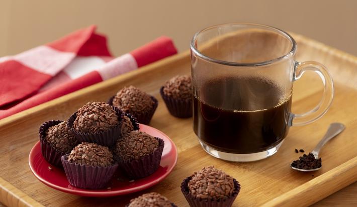 Fotografia em tons de marrom em uma bandeja de madeira de cor marrom. No centro, uma xícara de café preto e ao lado, alguns brigadeiros de Nescafé e uma colher contendo pó de Nescafé. Ao fundo, um pano vermelho.