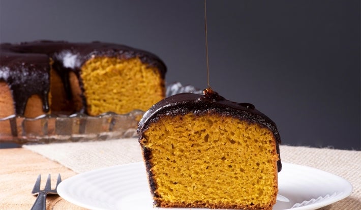 Um pedaço de bolo de cenoura com cobertura de chocolate brilhante está servido em um prato branco. A fatia está em destaque, e por cima dela há um fio de calda sendo derramado. Ao fundo, aparece o restante do bolo, também coberto com chocolate.